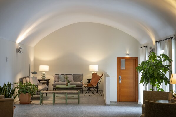 Una sala moderna con sofá, sillones, una mesa de centro y una mesa de comedor. Hay un ventanal con cortinas, una puerta con un número y una planta en maceta.
