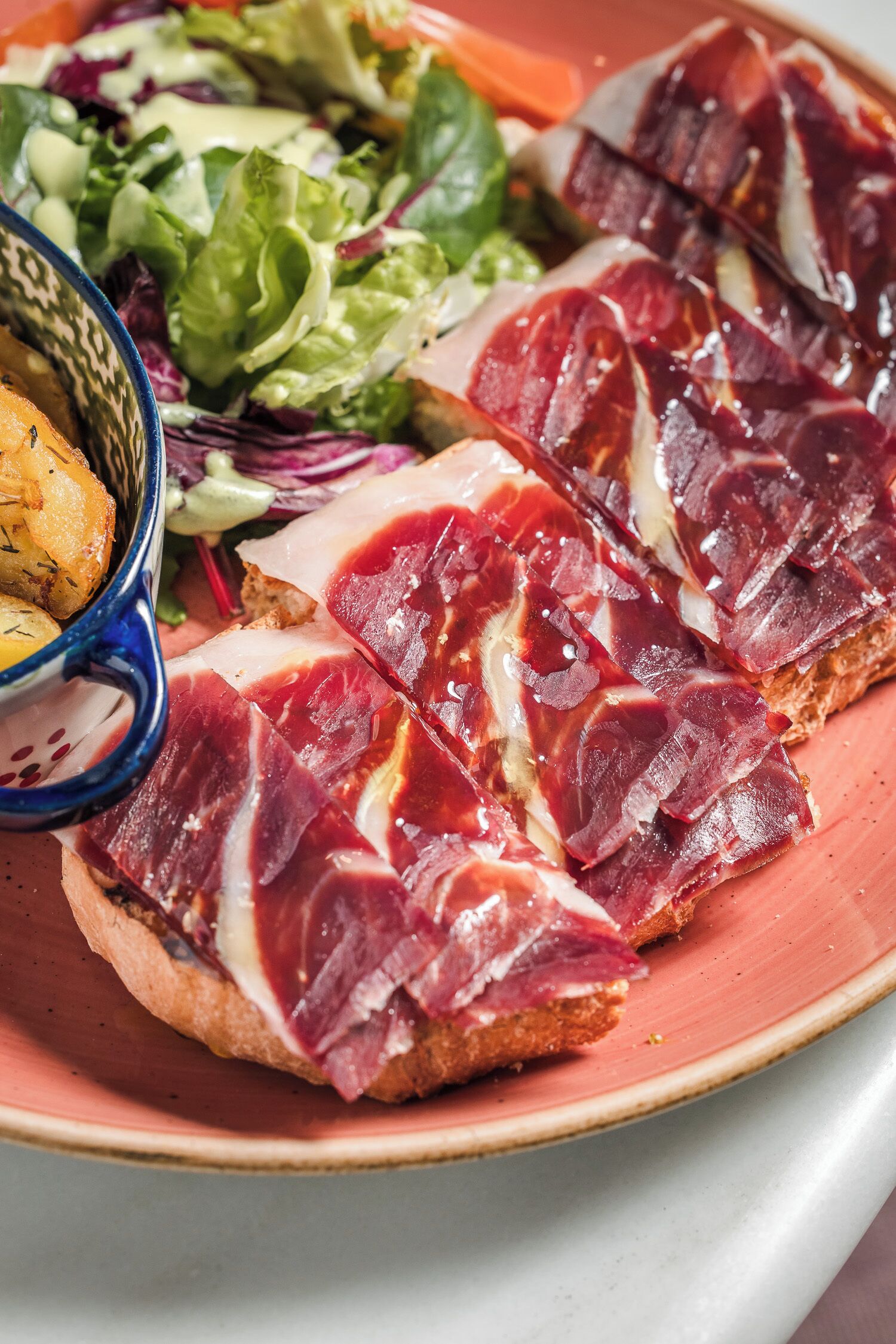 Un plato con carne curada en rodajas, un bol de patatas y una ensalada.