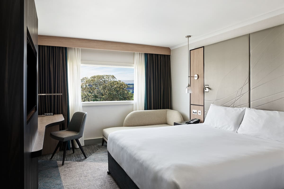 Une chambre d’hôtel avec un grand lit, un bureau, une chaise, une télévision et une fenêtre donnant sur des arbres et des immeubles.
