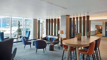 A modern hotel lobby with a large seating area, a wooden table, and a view of the street through large windows.