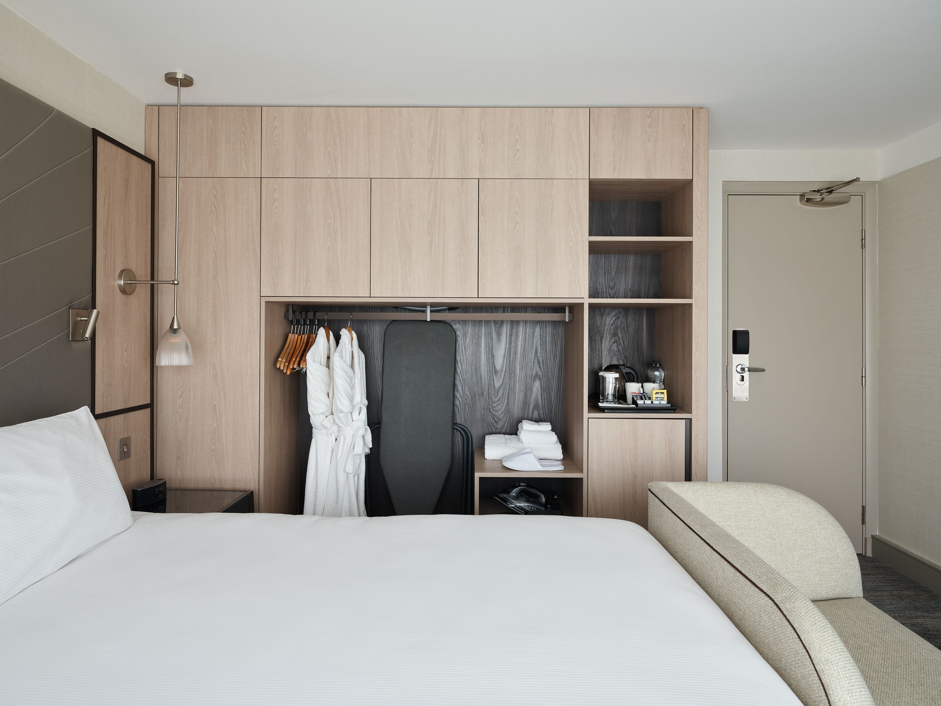 Une chambre d’hôtel moderne dotée d’un grand lit, d’un coin salon, d’une armoire avec plusieurs compartiments, ainsi que d’une petite table avec une bouilloire et des tasses.