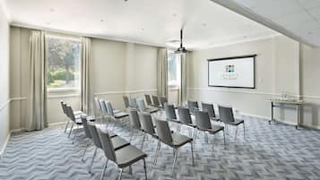 Un salón de conferencias con filas de sillas, una pantalla de proyector y un amplio ventanal con cortinas.