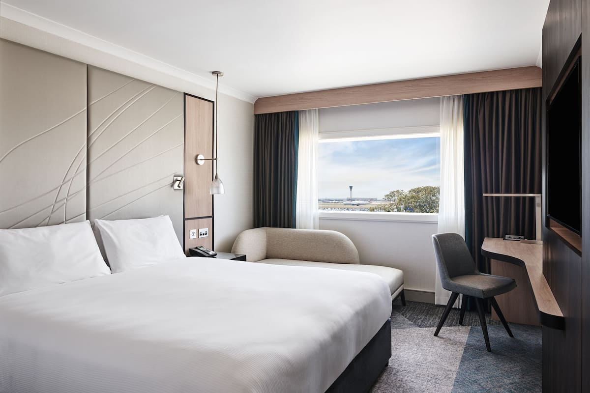 Une chambre d’hôtel avec un grand lit, un bureau, une chaise, une télévision et une fenêtre donnant sur un aéroport.