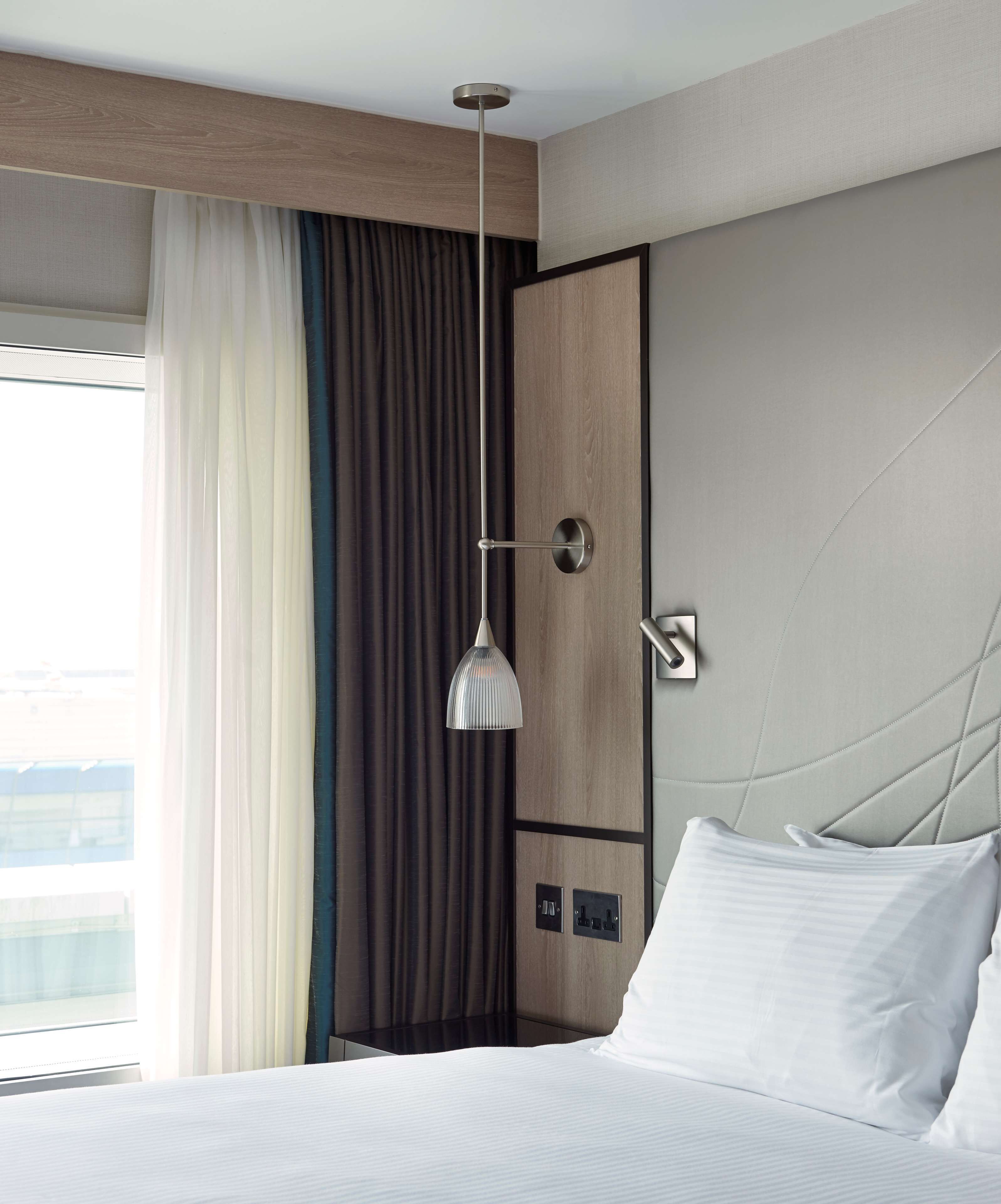 A hotel room with a bed, a hanging pendant light, a view of the city, and a large window with curtains.