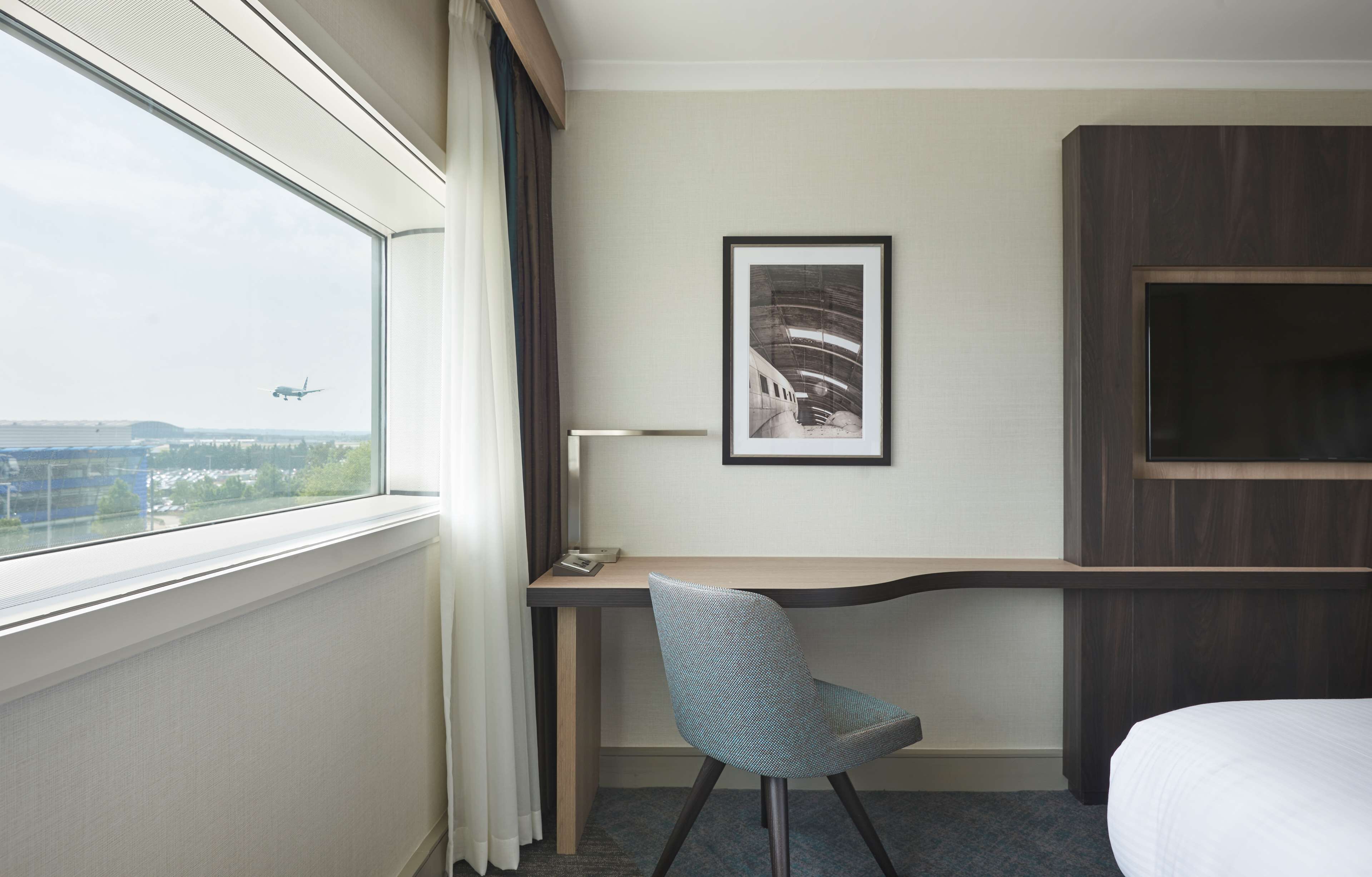 Hotellihuone, jossa on työpöytä, tuoli ja suuri ikkuna, josta avautuu näkymä lentokentälle.