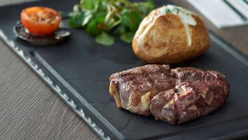 A plate with a steak, a baked potato, and a tomato on a dark slate.