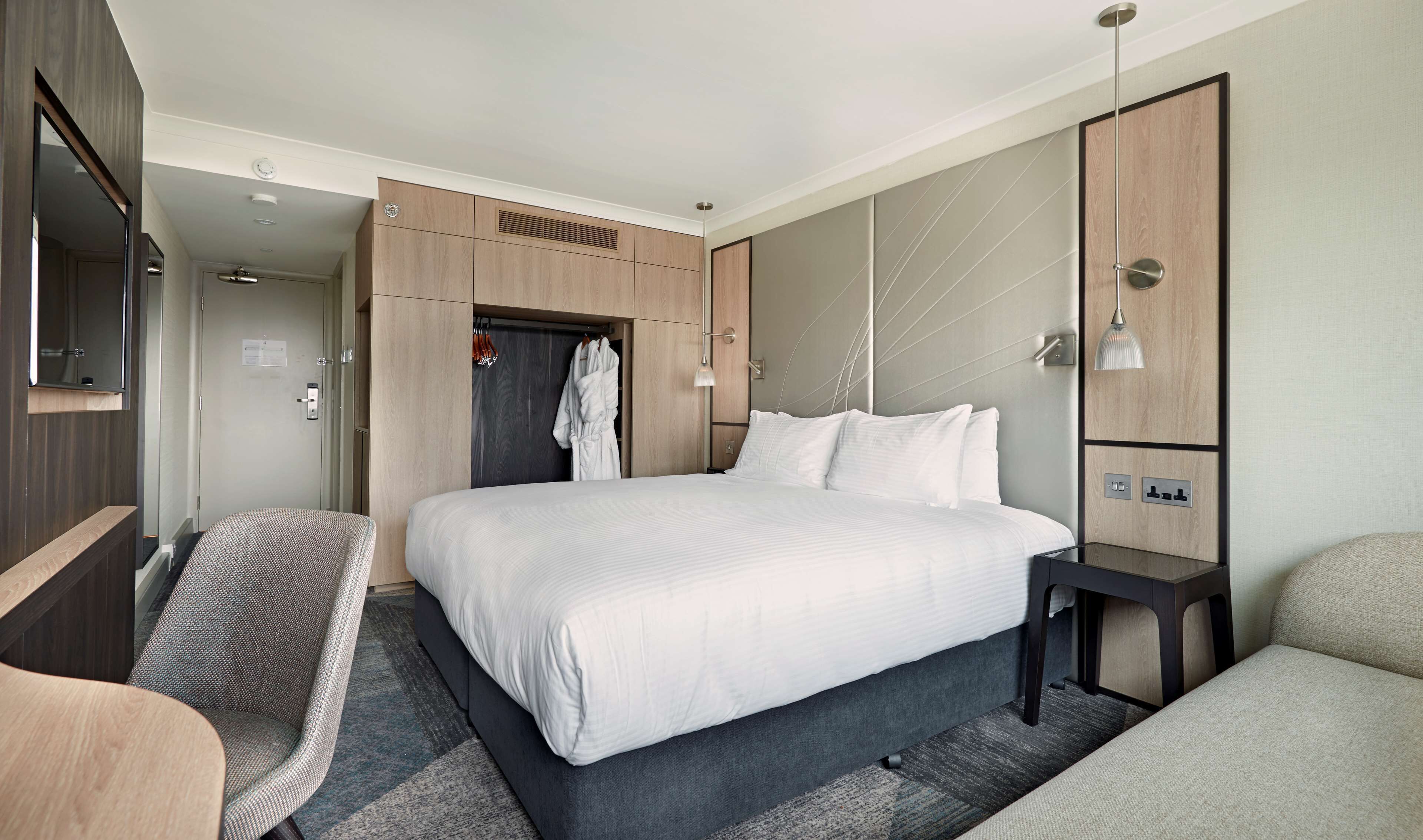 A modern hotel room with a large bed, a seating area with a chair, a wardrobe, and a mirror.