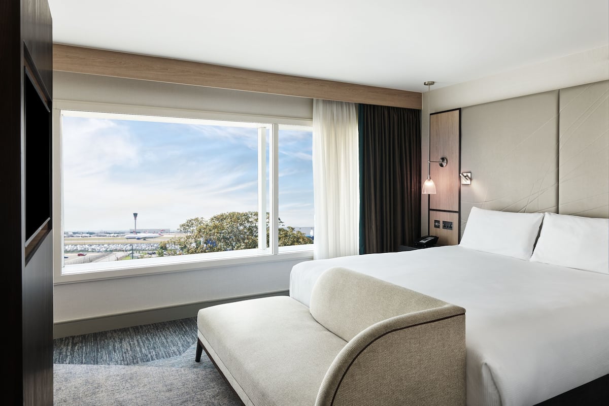 Une chambre d’hôtel avec un grand lit, une table de chevet, une lampe, une vue sur un aéroport et une fenêtre avec des rideaux.