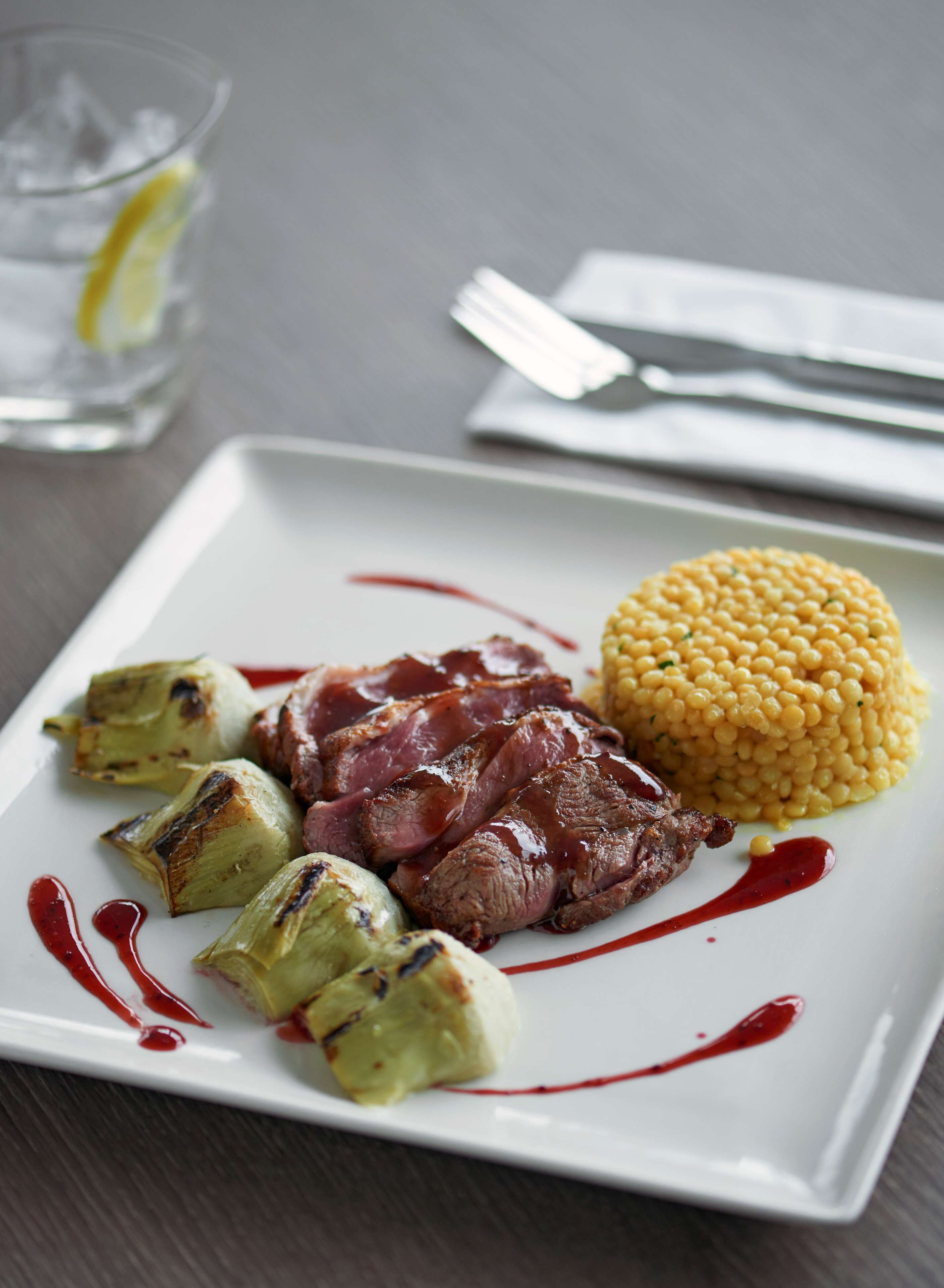A plate with grilled artichoke hearts, a slice of meat, and a mound of yellow corn kernels, all on a white plate with red sauce drizzles.