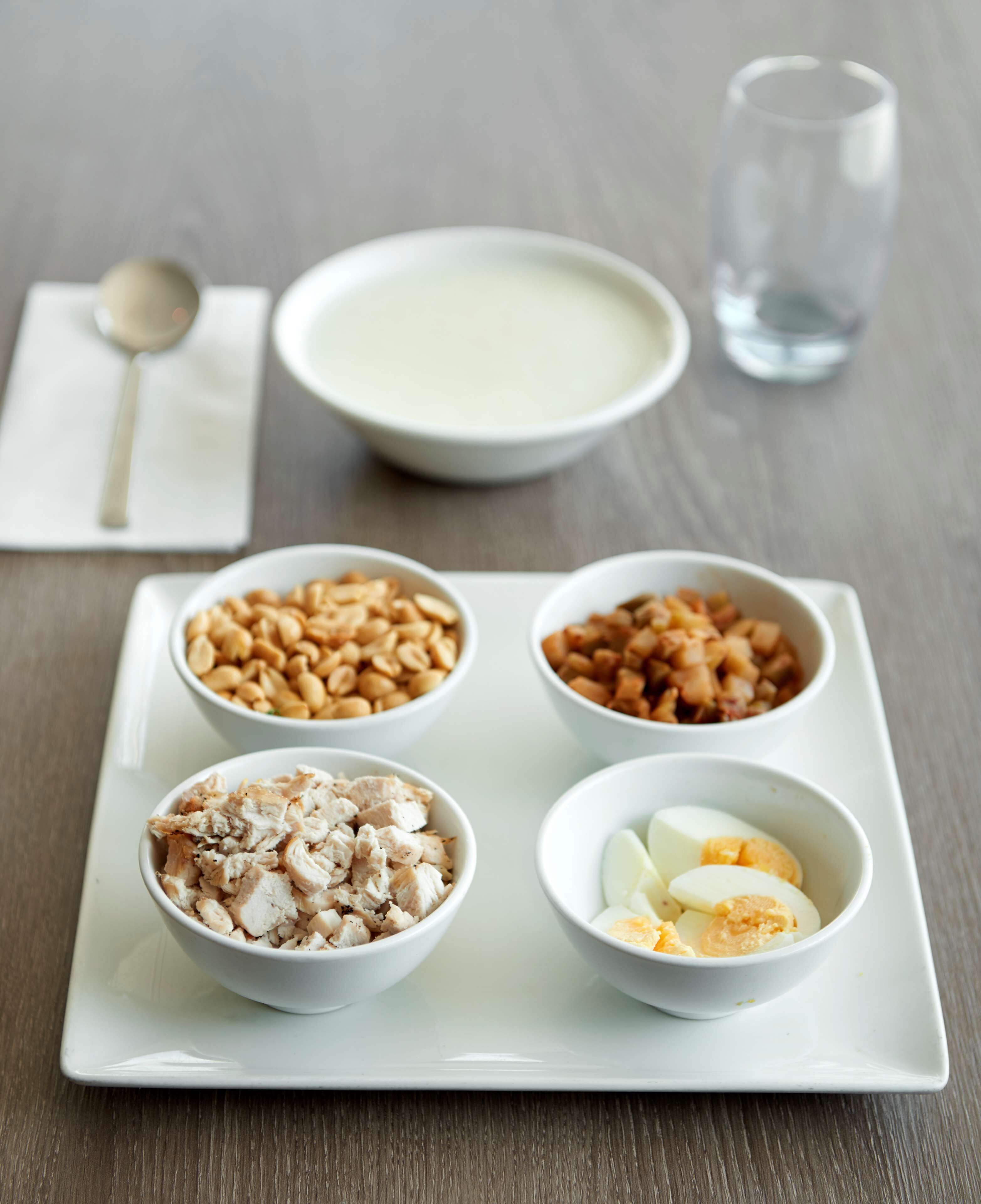 A white plate with four bowls of food: nuts, chicken, boiled eggs, and a bowl of yogurt.