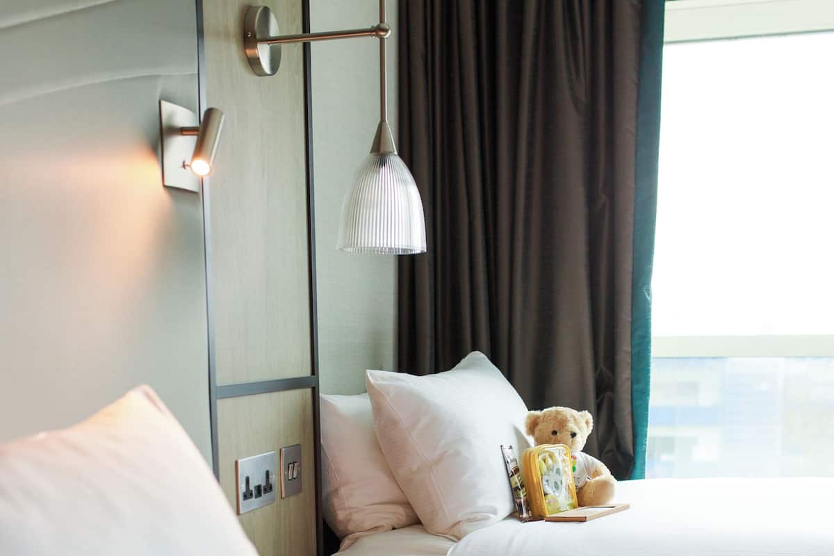 Une chambre d’hôtel avec deux lits, une lampe de chevet, un ours en peluche et une fenêtre avec des rideaux.