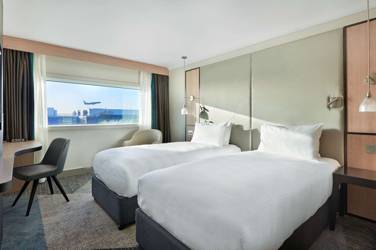 Une chambre d’hôtel avec deux lits, un bureau et une chaise. Il y a une fenêtre donnant sur des immeubles et un avion.