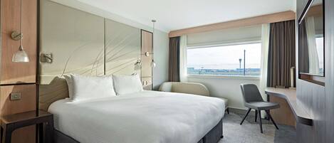 Habitación de hotel moderna con una cama grande, un escritorio y vista al aeropuerto.