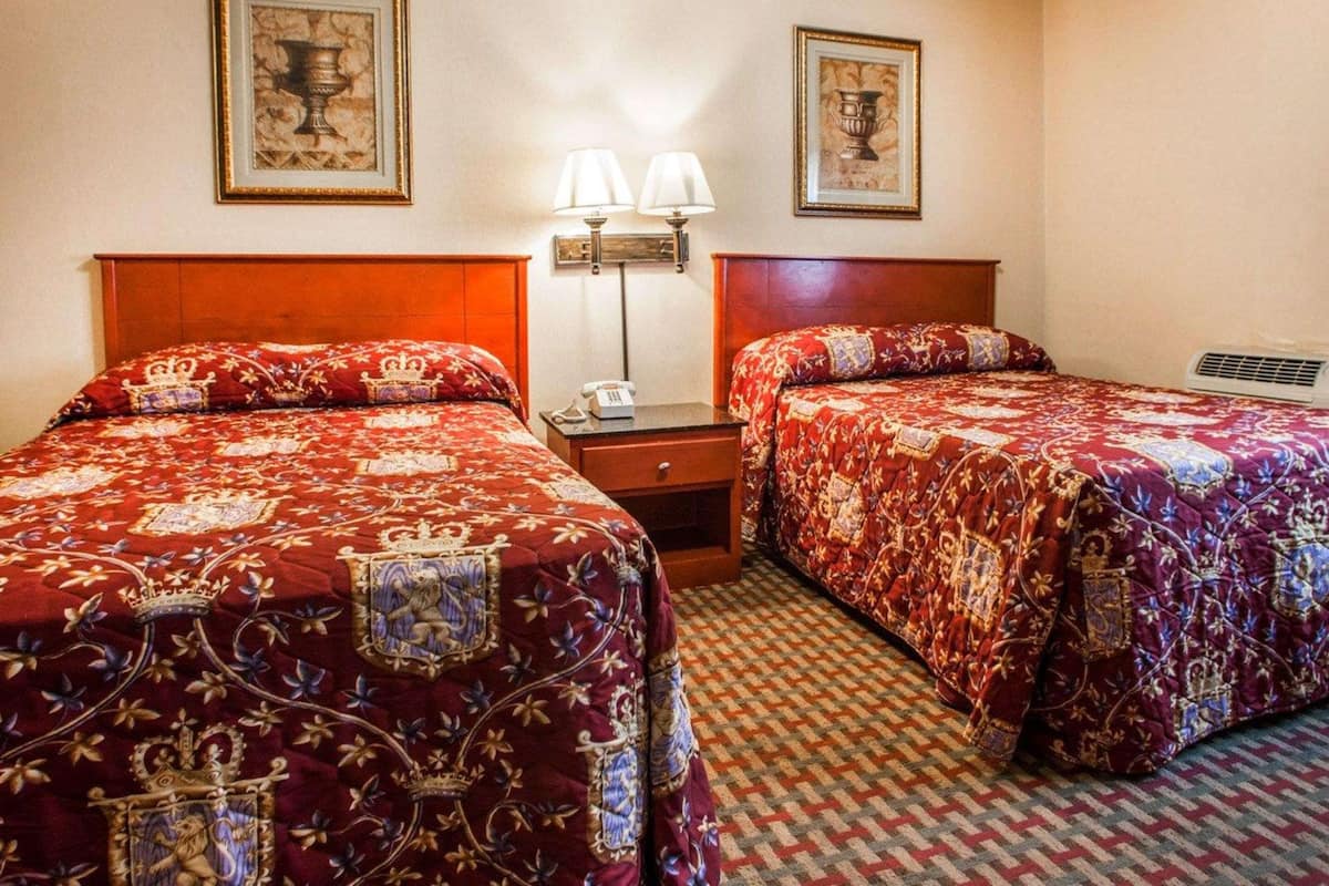 Una camera d'hotel con due letti, ognuno con una coperta rossa decorata con un'emblema di leone, un comodino con una lampada e un quadro incorniciato sopra il letto.
