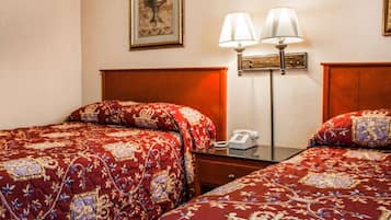 A hotel room with two beds, each with a red patterned bedspread, a wooden headboard, a bedside table with a lamp, and a framed picture on the wall.