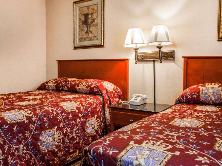 A hotel room with two beds, each with a red patterned bedspread, a wooden headboard, a bedside table with a lamp, and a framed picture on the wall.