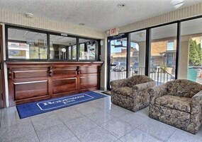 Uma área de recepção com uma mesa de madeira, duas poltronas estampadas e um tapete azul com o nome do hotel.