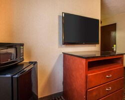 Quarto de hotel compacto com micro-ondas, mini-geladeira, cômoda de madeira com gavetas e TV de tela plana na parede.