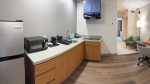 Cocina compacta con refrigerador, microondas y cafetera, un escritorio de madera con una silla, y vista hacia una sala de estar.