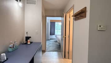 A hallway leading to a bedroom with a bed, a desk with a coffee machine, and a window with curtains.