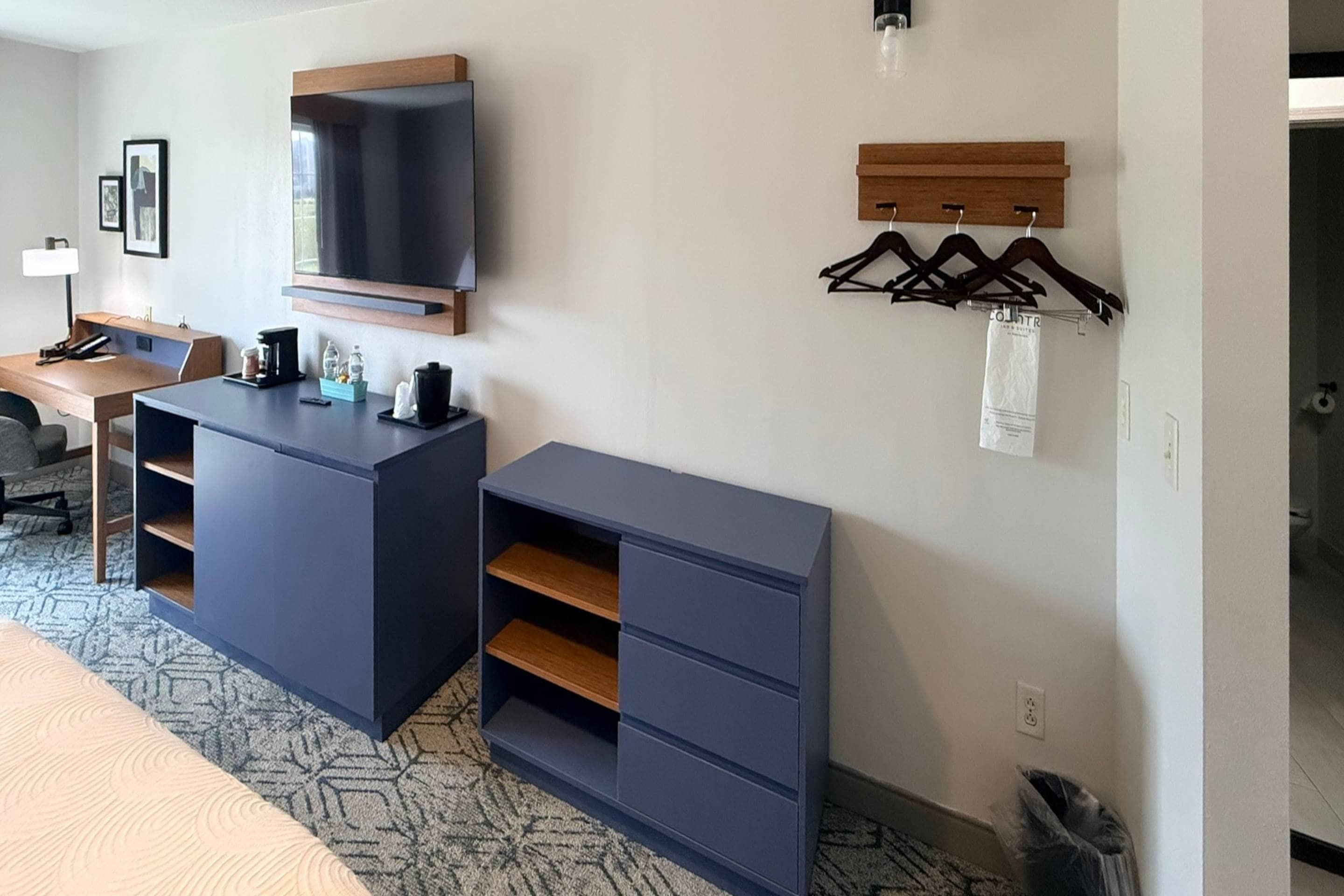 A hotel room with a desk, a flat-screen TV mounted on the wall, a coat rack with hangers, and a trash bin.