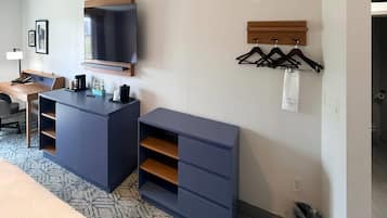 A hotel room with a desk, a flat-screen TV mounted on the wall, a coat rack with hangers, and a trash bin.