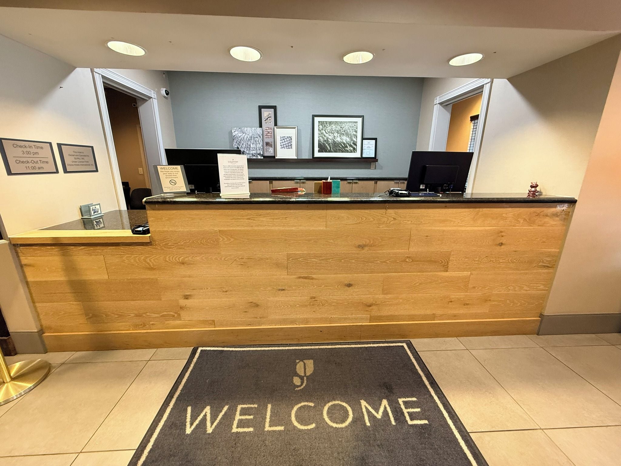 A reception desk with a 'WELCOME' mat, framed pictures on the wall, and a check-in/out schedule.