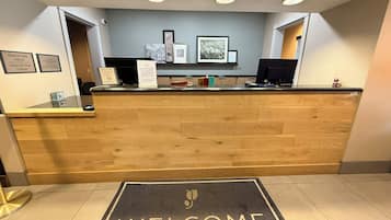 A reception desk with a 'WELCOME' mat, framed pictures on the wall, and a check-in/out schedule.