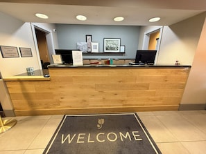 Un comptoir d’accueil avec un tapis où est inscrit « BIENVENUE », des photos encadrées accrochées au mur et un tableau indiquant les heures d’arrivée et de départ.