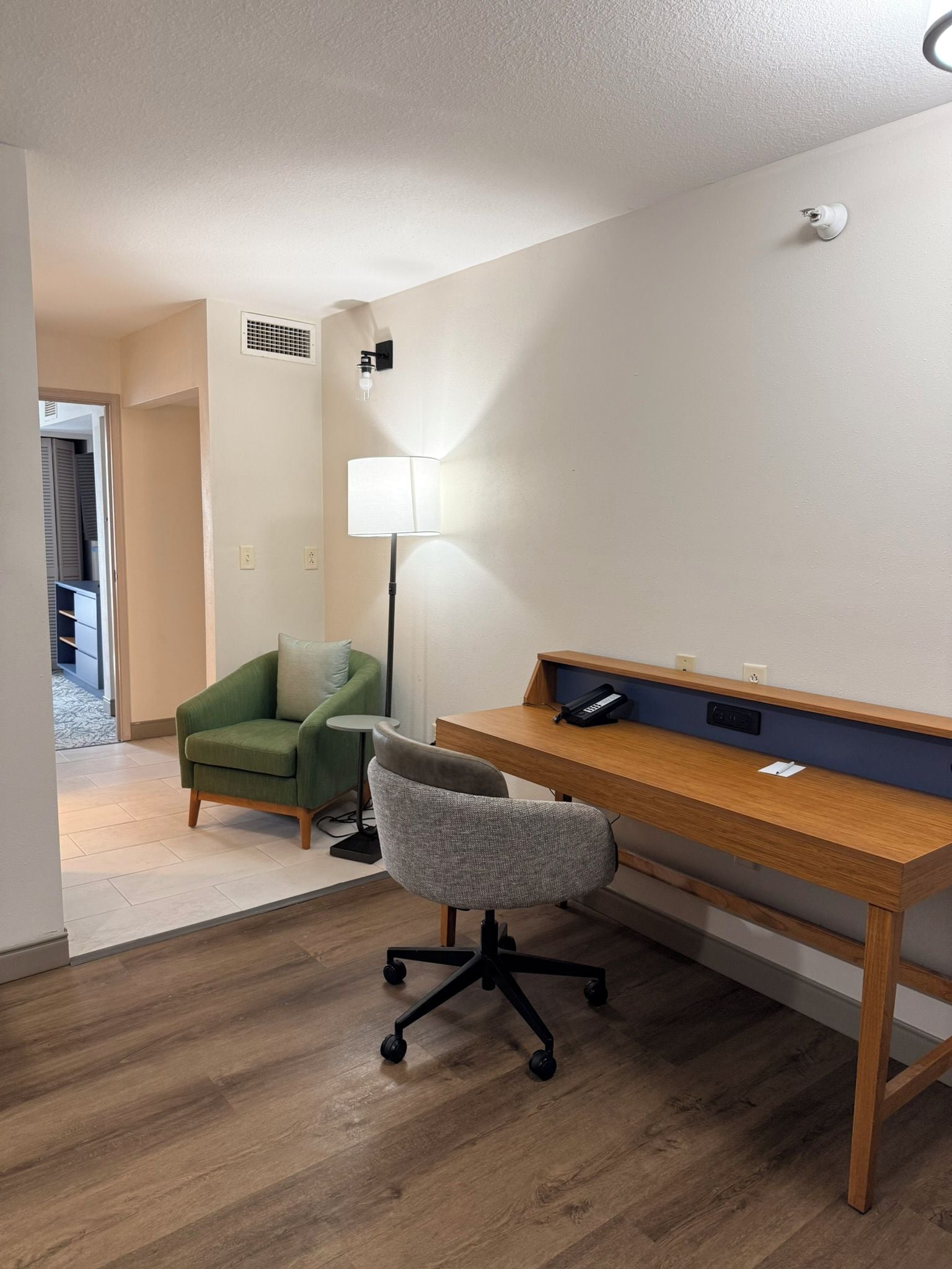 A room with a wooden desk, a green armchair, a grey office chair, a floor lamp, and a small table with a phone.