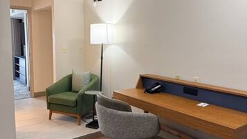 A room with a wooden desk, a green armchair, a grey office chair, a floor lamp, and a small table with a phone.