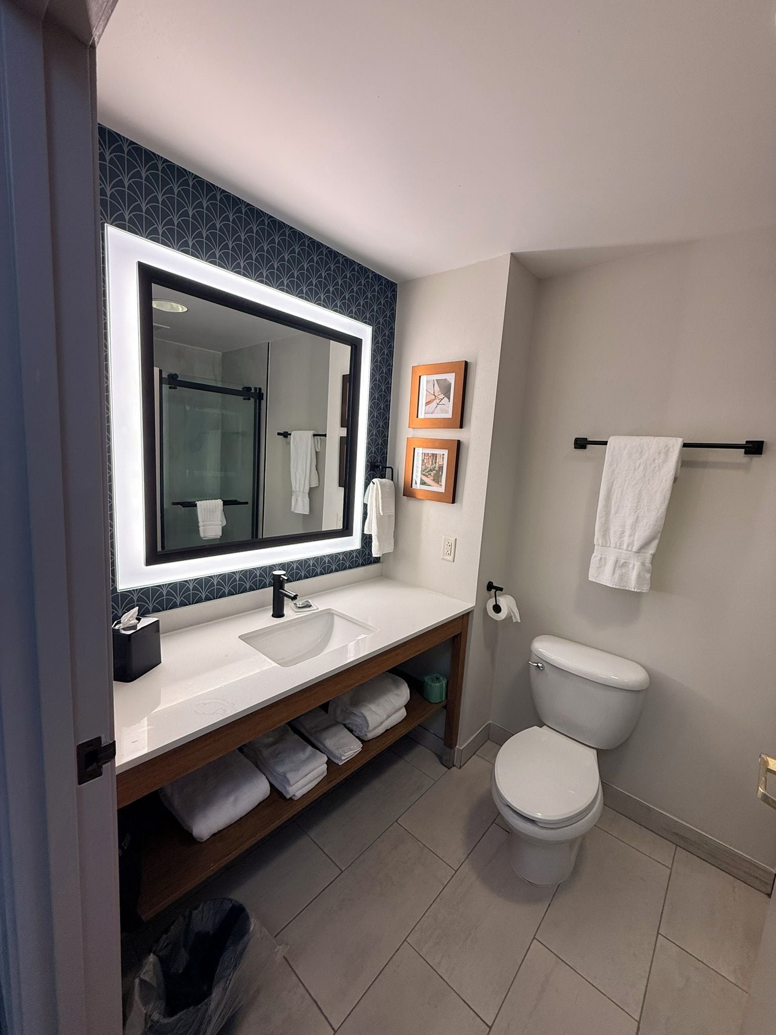 A modern bathroom with a large mirror, white sink, and toilet. There are towels and a shelf with folded towels.