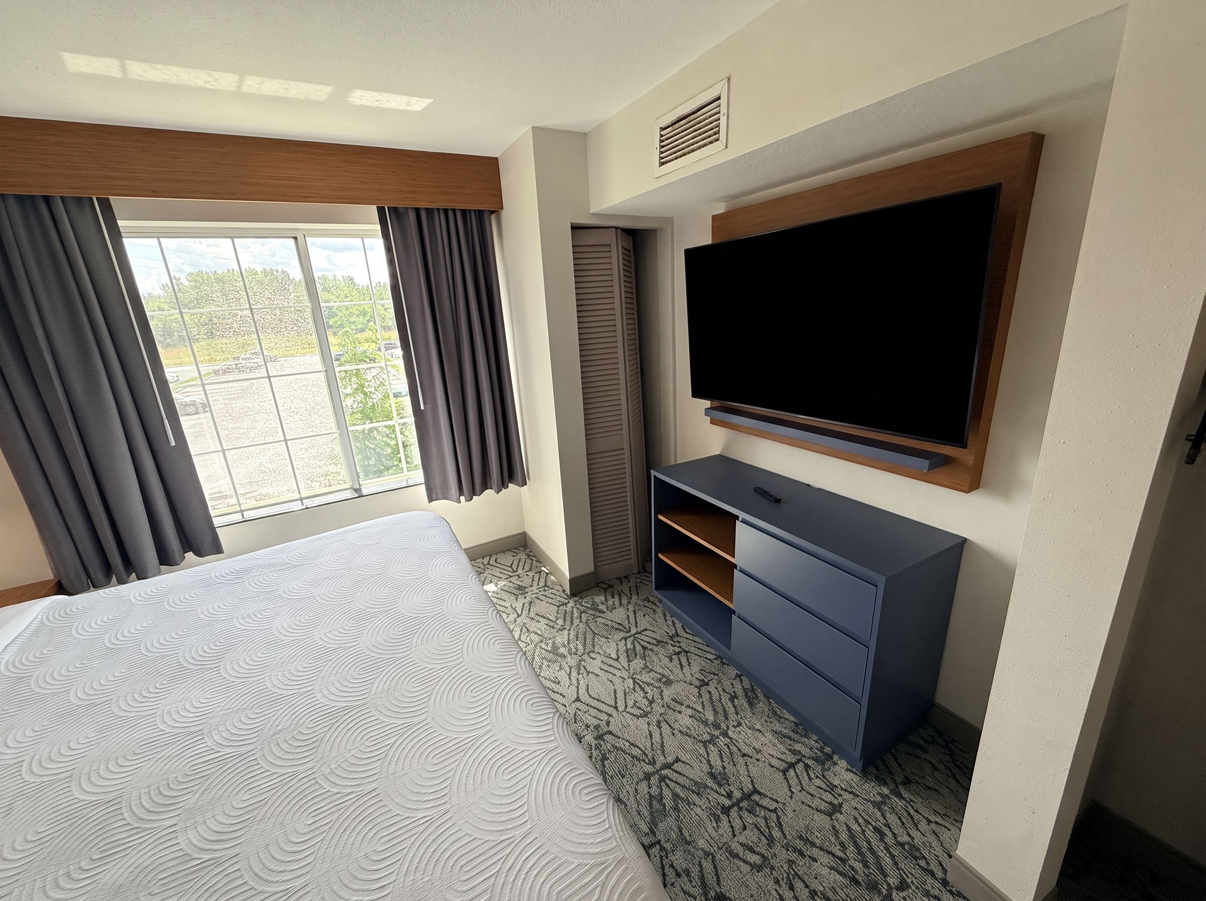 Quarto de hotel com cama, televisão de tela plana na parede, escrivaninha e janela com cortinas.
