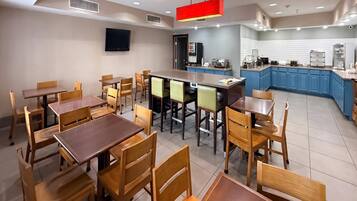 A hotel dining area with multiple wooden tables and chairs, a kitchen area with blue cabinets, and a flat-screen TV mounted on the wall.