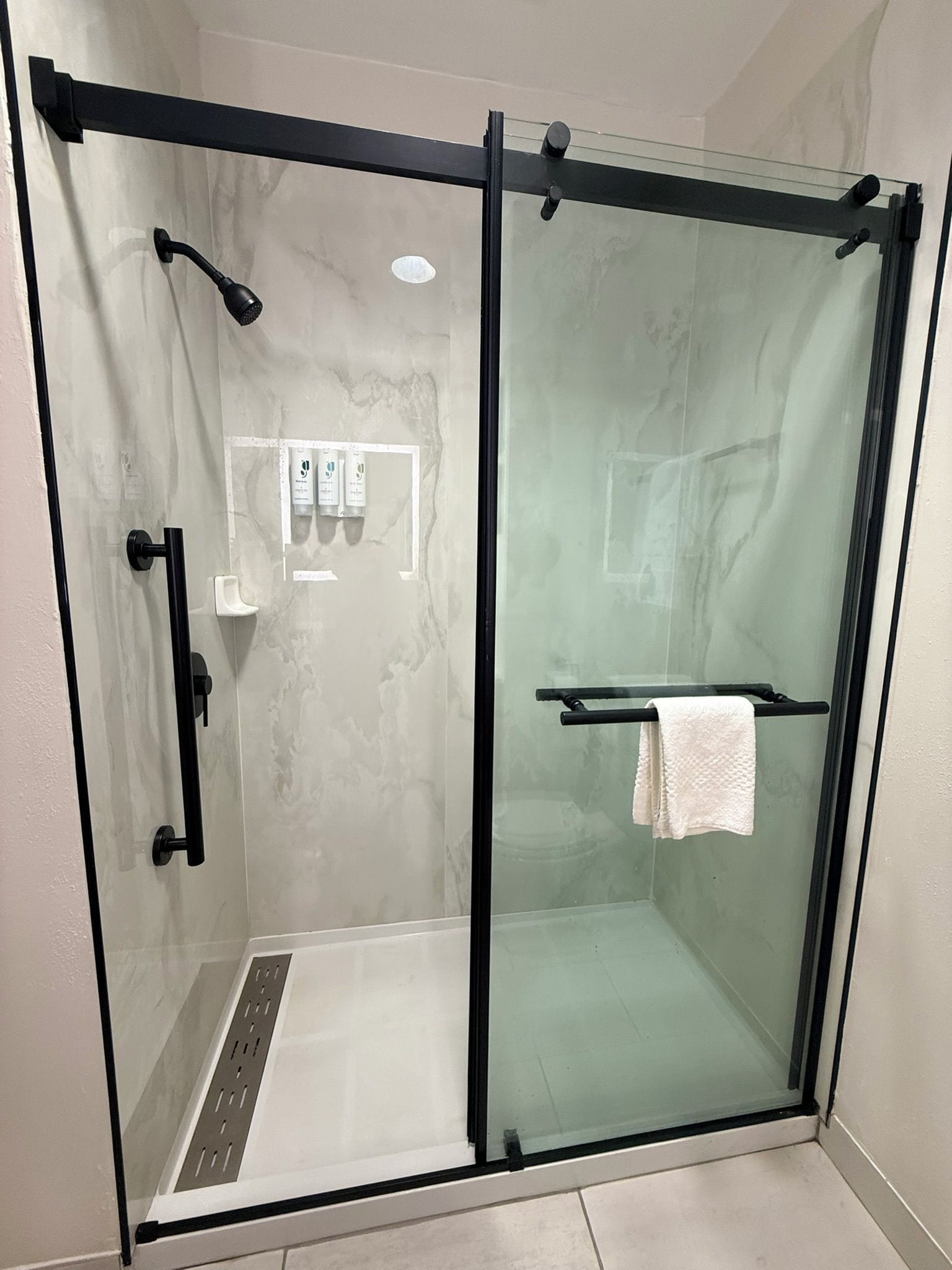 A glass-enclosed shower stall with a black frame, a white towel on a rack, and a small shelf with toiletries.