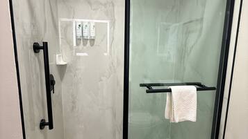 A glass-enclosed shower stall with a black frame, a white towel on a rack, and a small shelf with toiletries.