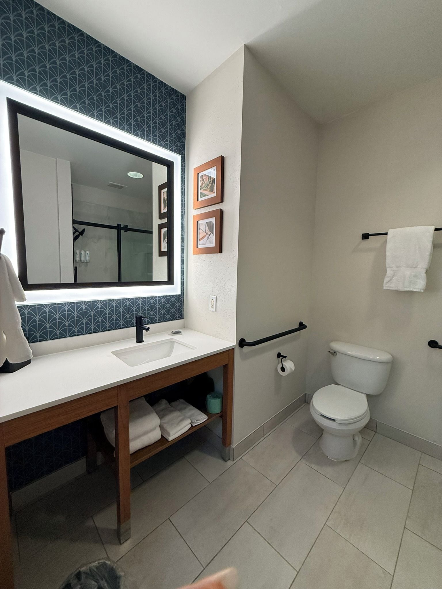 Un baño moderno con un espejo grande, un lavamanos blanco y un inodoro. Hay cuadros en la pared y un perchero para toallas.