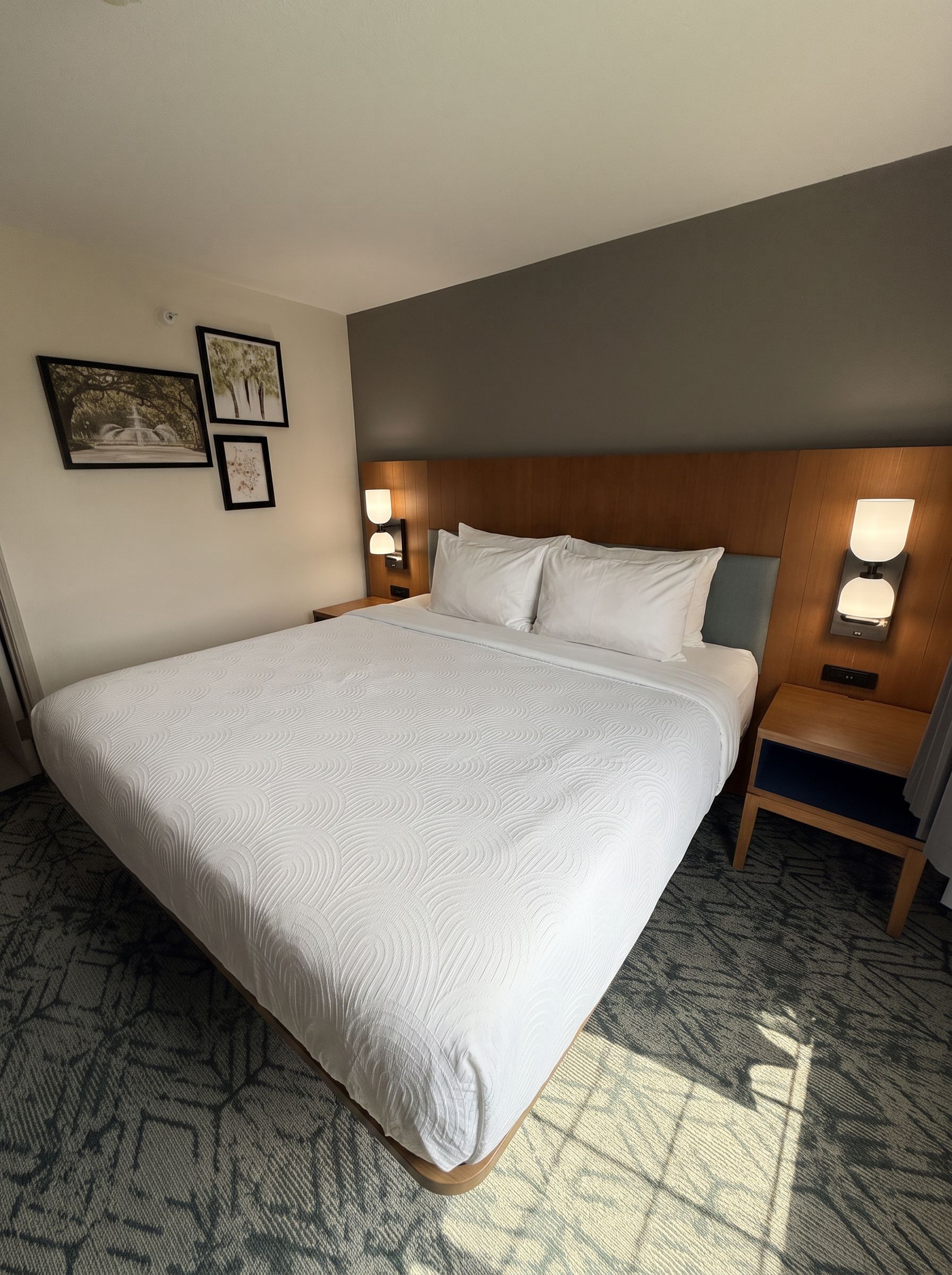 Quarto de hotel com uma cama grande, duas mesas de cabeceira e luminárias fixadas na parede.