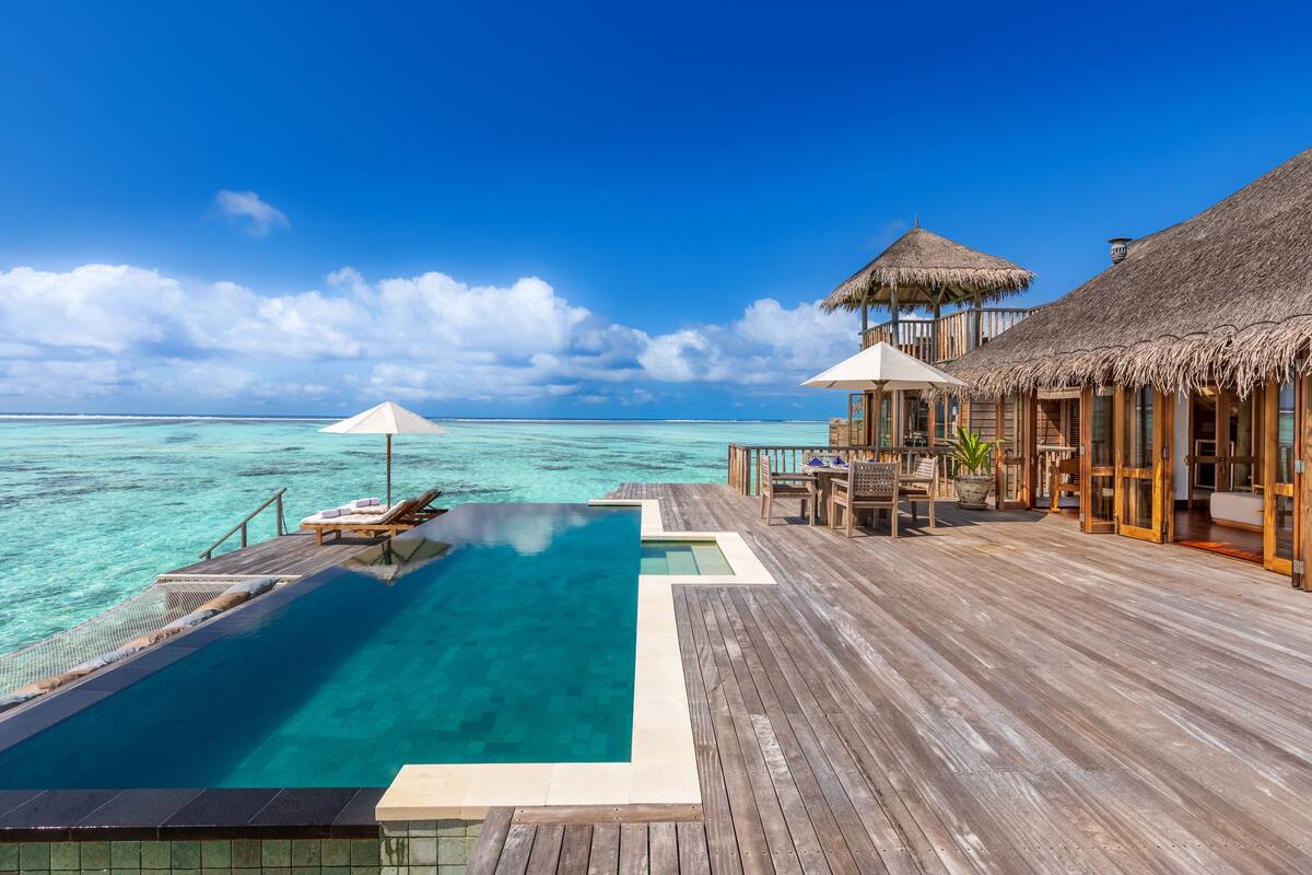 Ein Infinity-Pool mit klarem, blauem Wasser, einer Holzterrasse und Bungalows auf Stelzen mit Reetdächern.