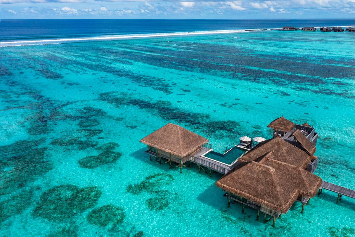 Eine Vogelperspektive zeigt Bungalows auf Stelzen mit strohgedeckten Dächern, verbunden durch Gehwege, umgeben von klarem türkisblauem Wasser und Korallenriffen.