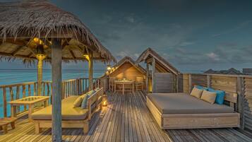 Una terraza de madera con una estructura de techo de paja, una cama de madera y una mesa con sillas.