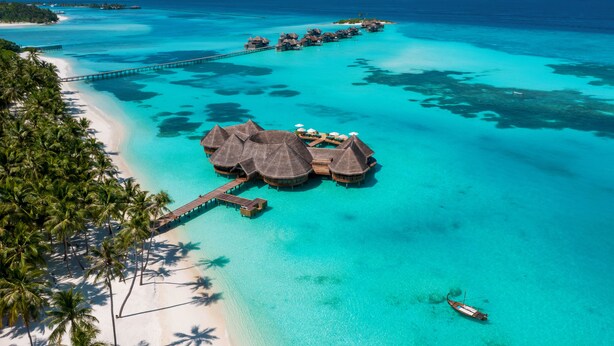 Een resort met bungalows op het water, voorzien van rieten daken, een pier en helder turquoise water.
