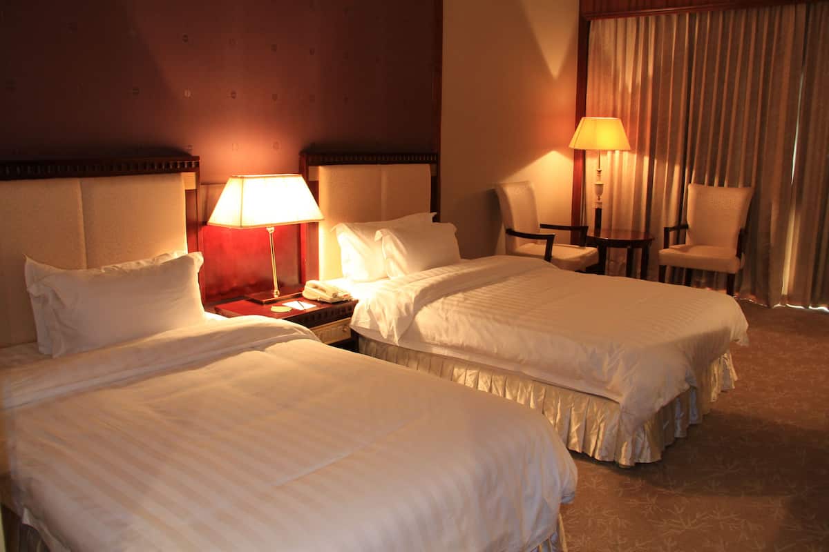 Quarto de hotel com duas camas, mesa de cabeceira, abajur, cadeira e cortina.