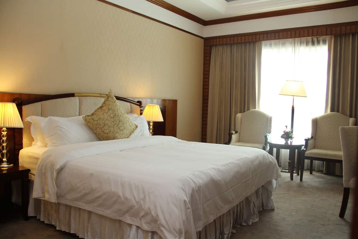 Quarto de hotel com uma cama grande, duas luminárias de cabeceira, uma mesinha com uma cadeira e uma janela com cortinas.