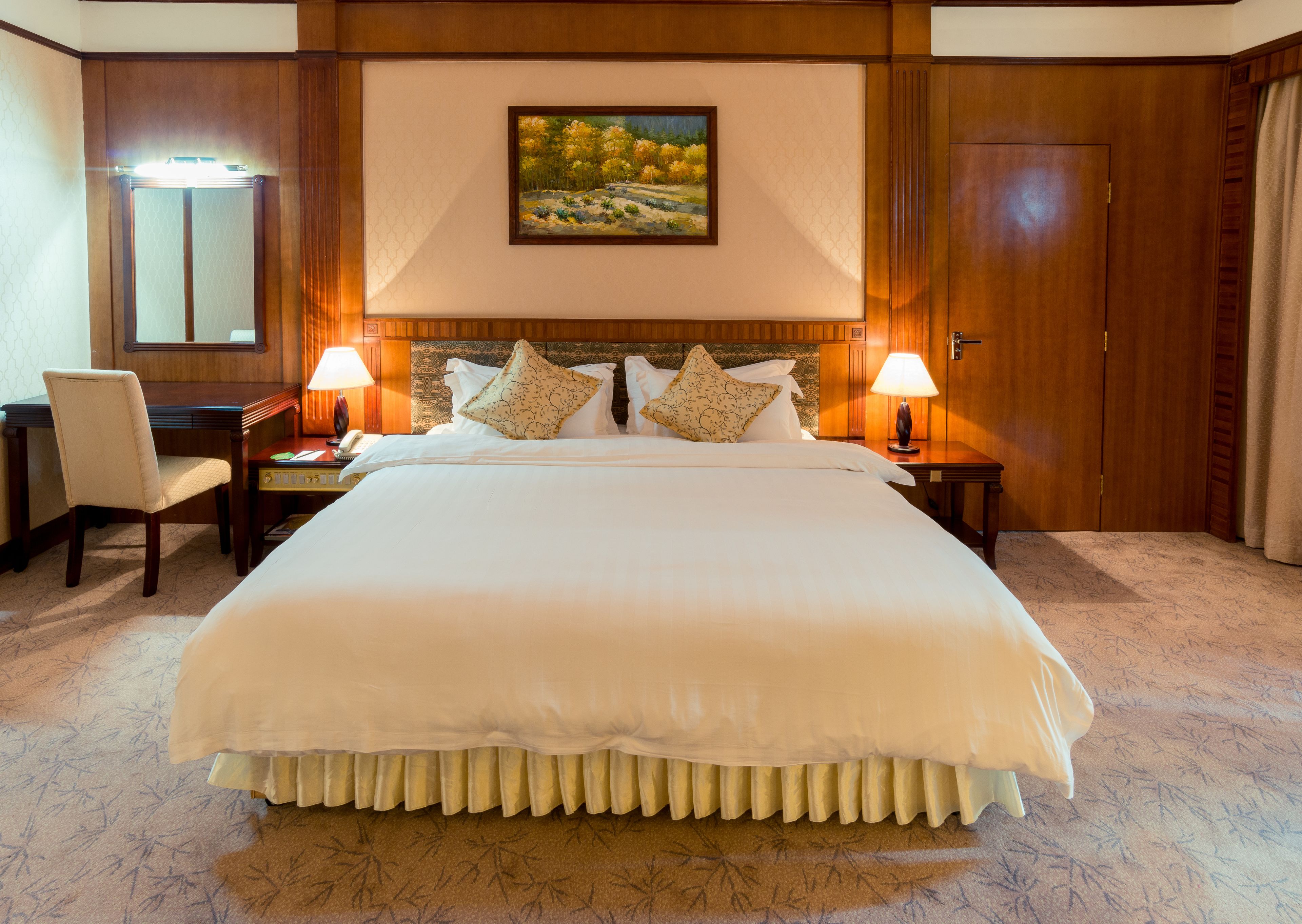 A large bed with a white duvet, two bedside tables with lamps, a chair, a painting on the wall, and a wooden door.