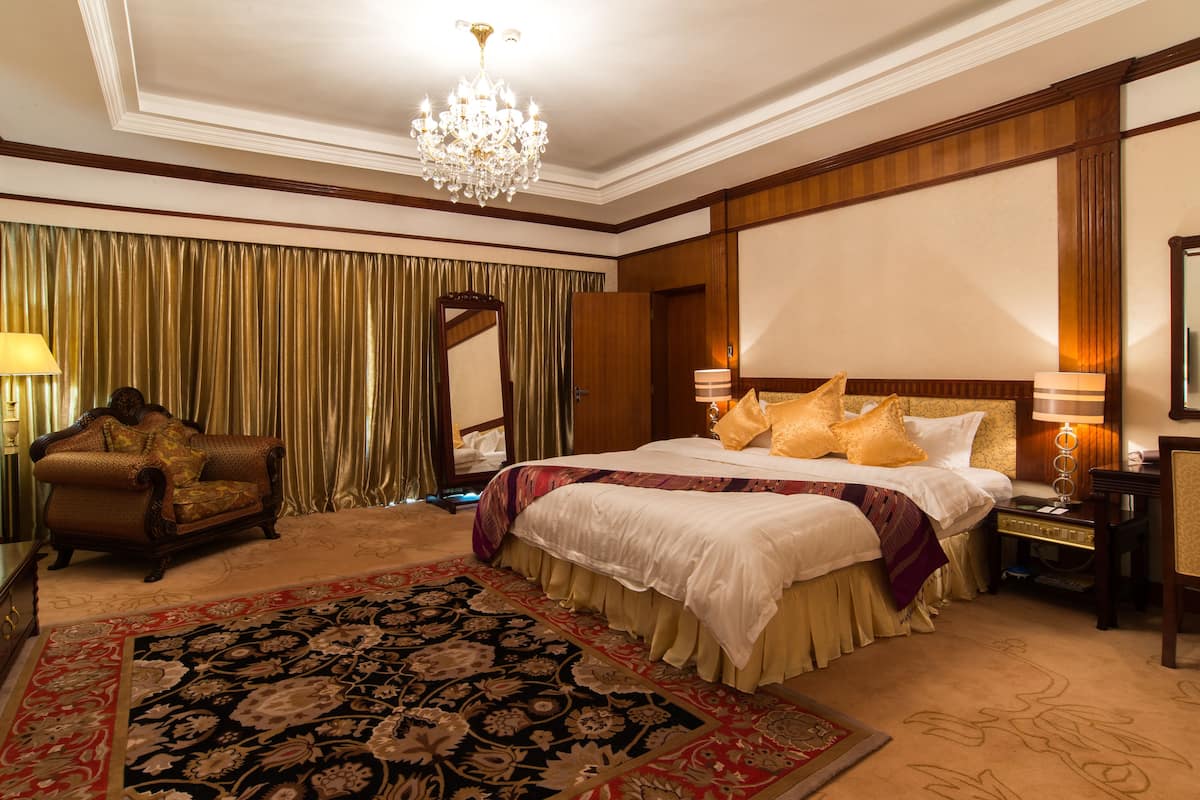 Um quarto de hotel espaçoso com uma cama grande, um espaço de estar com uma poltrona, um candelabro e um espelho grande.