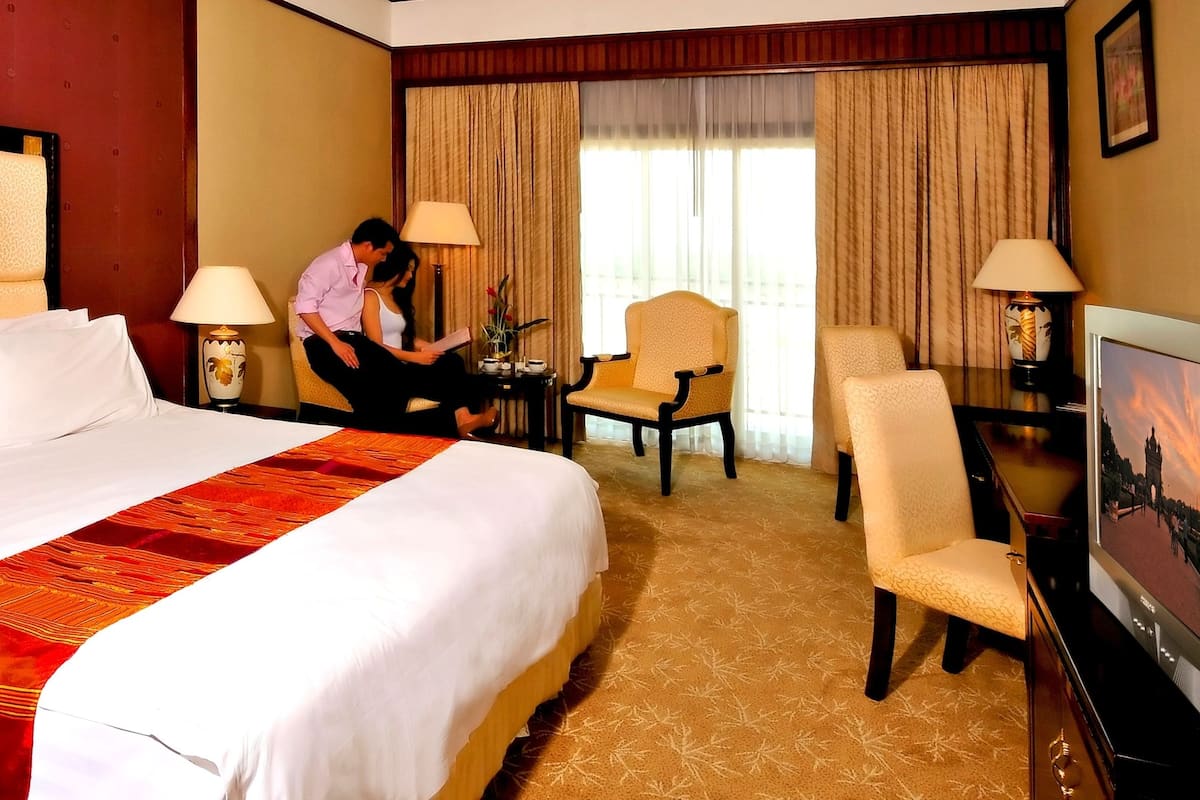 Quarto de hotel com uma cama grande, uma escrivaninha, duas cadeiras, uma televisão e uma janela com cortinas.