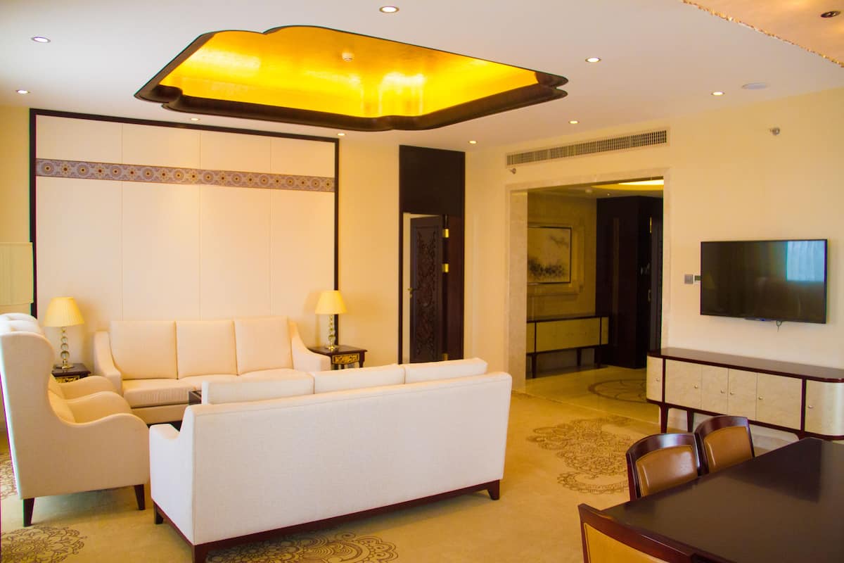 Quarto de hotel com sofá, duas mesas laterais com luminárias, uma mesa de jantar com cadeiras, uma TV de tela plana na parede e um tapete estampado.