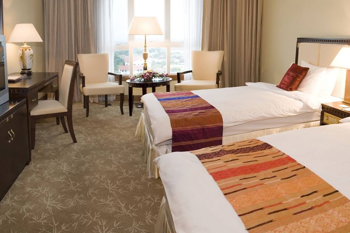 Quarto de hotel com duas camas, uma escrivaninha com televisão, uma cadeira, uma pequena mesa com abajur e vista para o exterior através de grandes janelas.