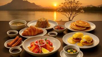 Une table dressée avec divers éléments pour le petit-déjeuner, notamment des croissants, des crêpes et des œufs Bénédicte, le tout disposé devant un magnifique panorama sur un lac au coucher du soleil.